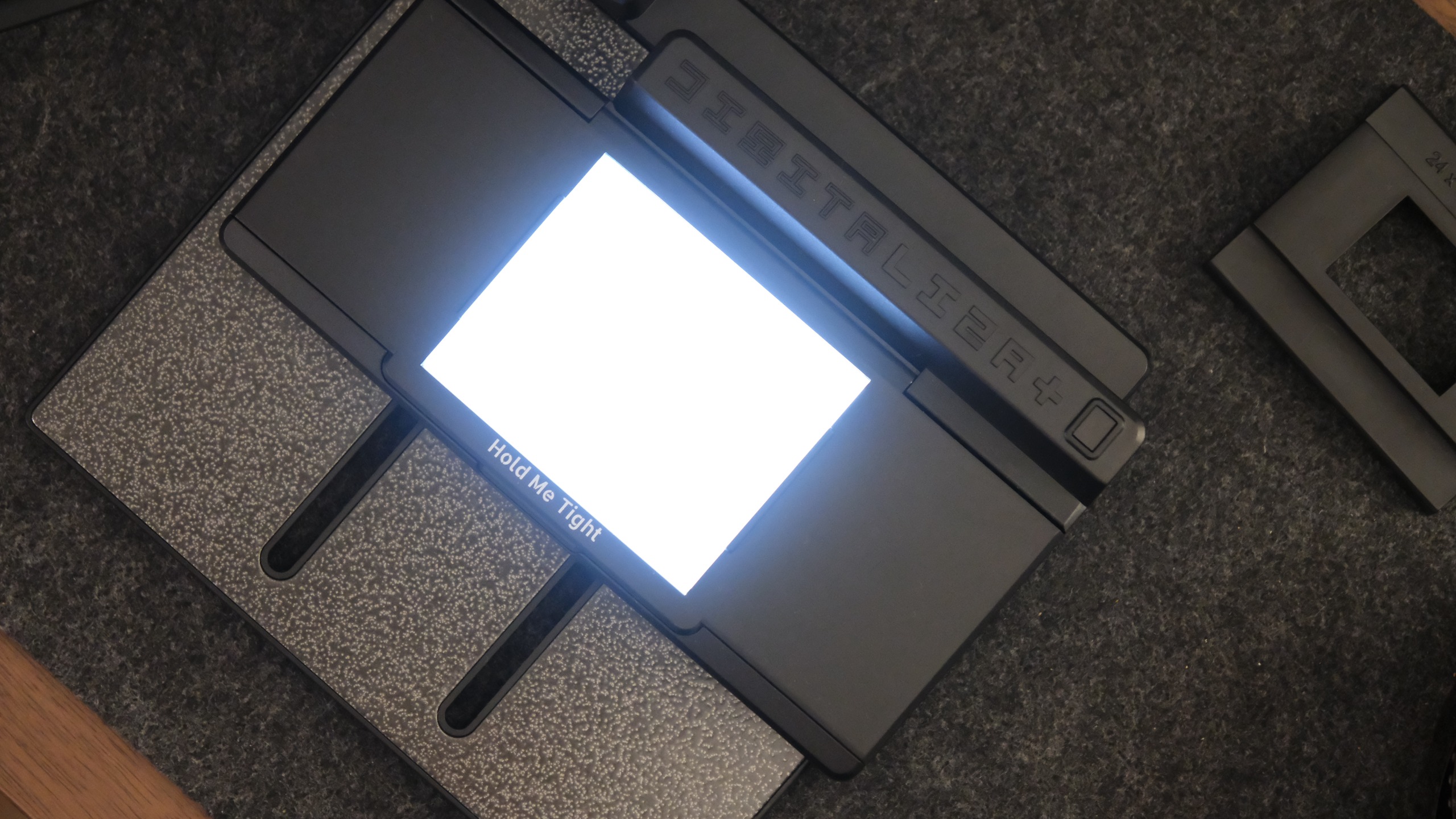 A top-down perspective of a black film digitizer base illuminating a bright white square of light from its center, with the text "Hold Me Tight" printed along the lower edge of the glowing window.