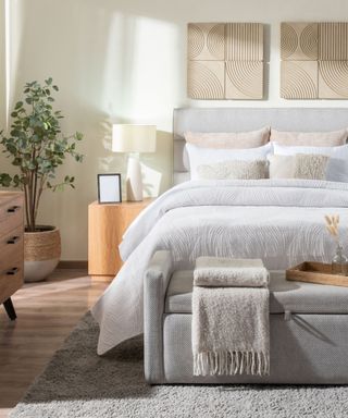 A bright bedroom with grey bed, storage ottoman with wooden tray and beige blanket and carpet, with wooden chest of drawers and bedside table, tall houseplant, white lamp, empty picture frame, off-white bedding, cushions and abstract wall art