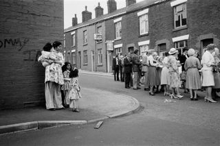 Asian mother and children stood on street corner in a residential street, looking at a queue of people opposite them