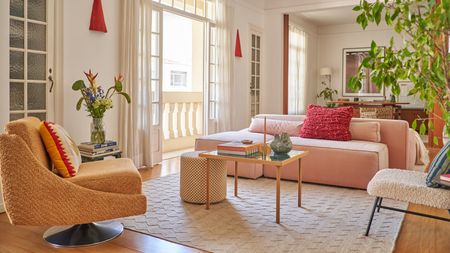 image of a bright living room that has a pink sofa and an orange accent chair across from it. There is a white rug on the floor and small wooden, square coffee table in front of the pink sofa.