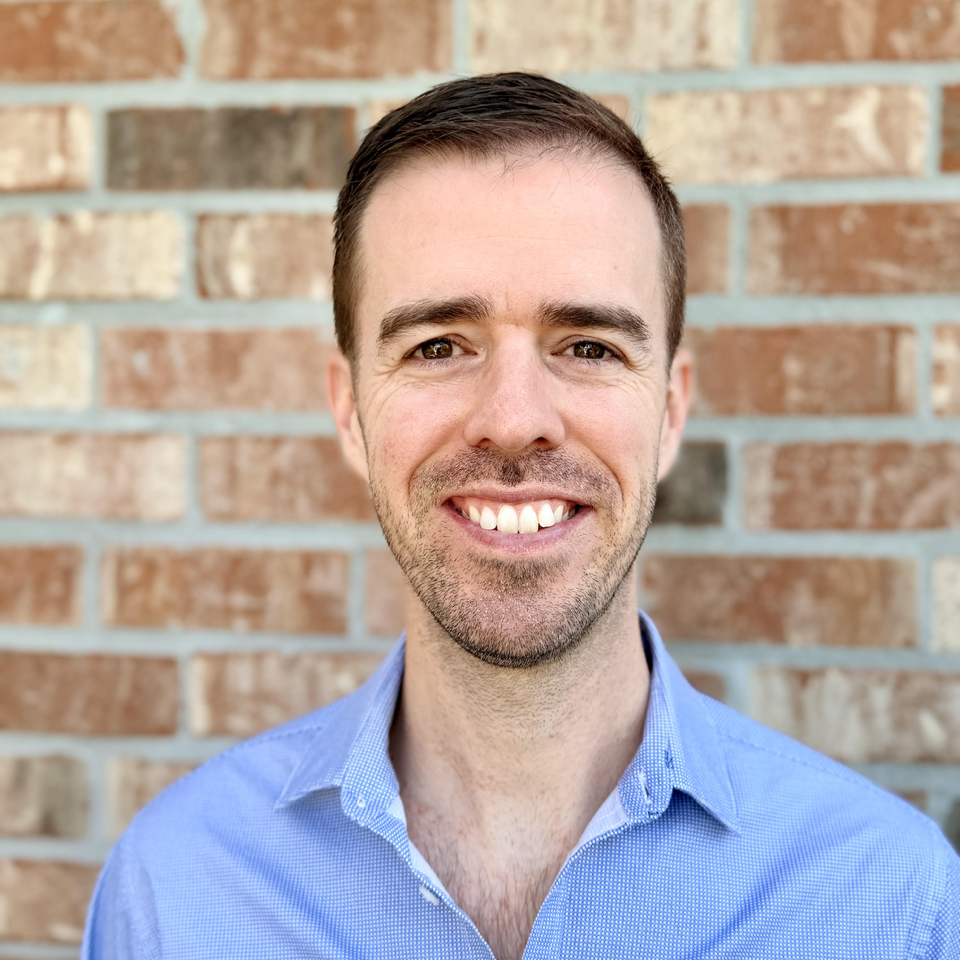 Headshot of man smiling in blue shirt against brick wall