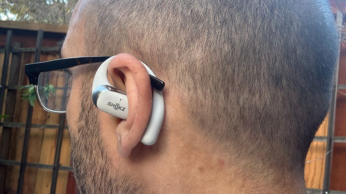 A self-photo of the author wearing a Shokz OpenFit 2 Plus earbud on his left ear, angled to show the hook behind his ear fitting next to his glasses arm.