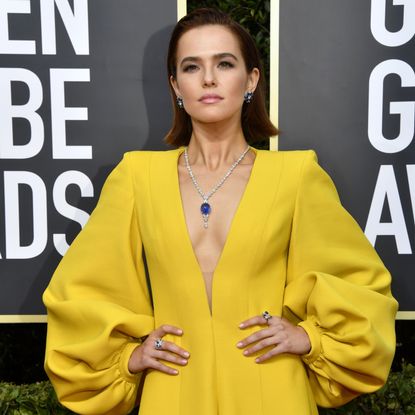 Zoey Deutch in a yellow jumpsuit with hands on hips wears a necklace with a large sapphire.