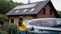 woman using EV charger in car stood in front of home with solar panels on roof