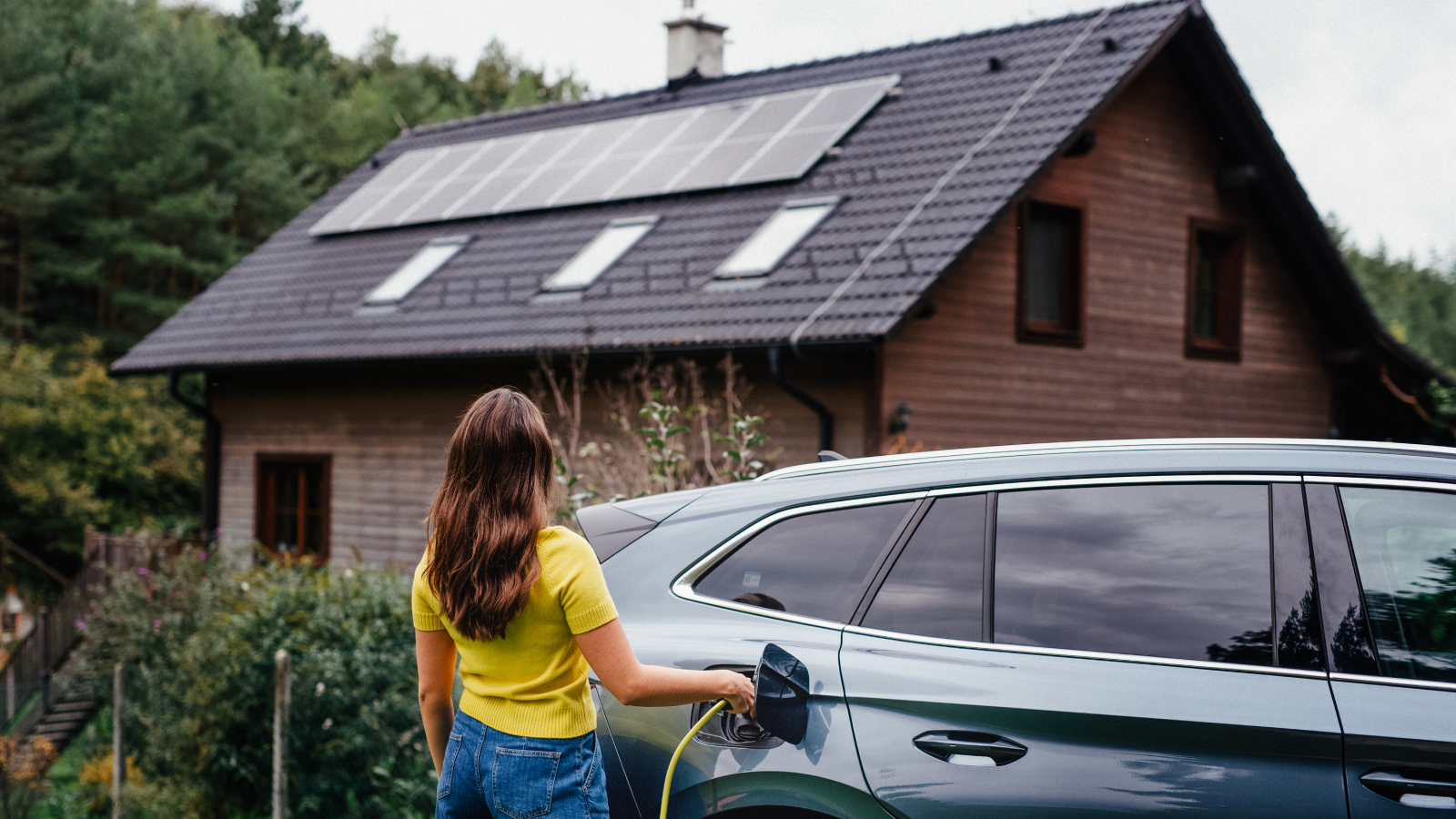 woman using EV charger in car stood in front of home with solar panels on roof