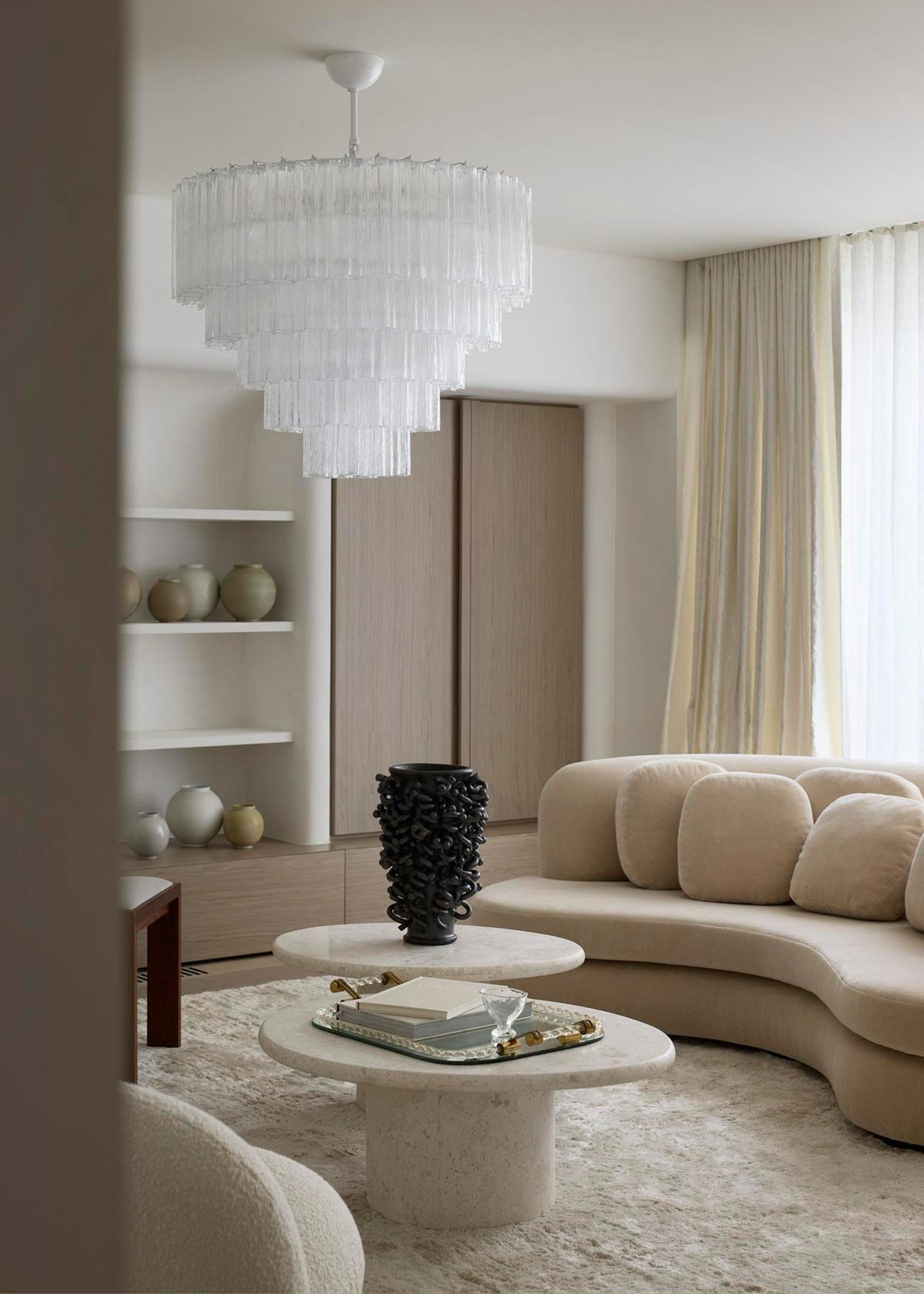 A minimalist living room with a mod chandelier, a curved sand beige couch with rounded square pillows, an area rug, a set of stone coffee tables with a tray and a black vase, and a wall of ceramics