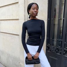Influencer @sylviemus_, wears white trousers with a black fitted top, gold statement earrings and a black leather clutch bag.