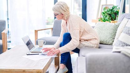 A retired woman sitting on her couch pays bills from her computer.