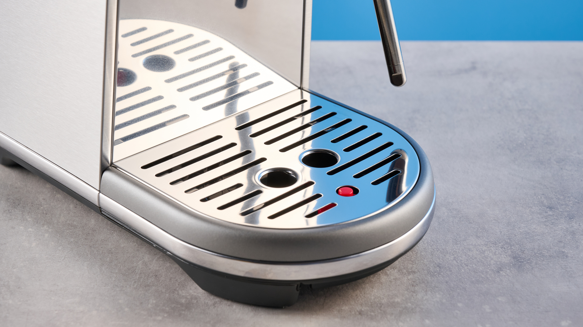 the breville bambino / sage bambino in silver photographed against a blue background with a wooden feature wall, showing the steam wand, group head, water tank, button controls, and espresso