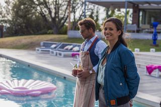 An image from Cannes Confidential showing Harry King (Jamie Bamber) and Camille Delmasse (Lucie Lucas) stand next to a swimming pool in the back garden of a large house. She has her head turned towards the camera and is smiling, he is facing to the left and wearing sunglasses and an apron, carrying two glasses of elaborate-looking drinks
