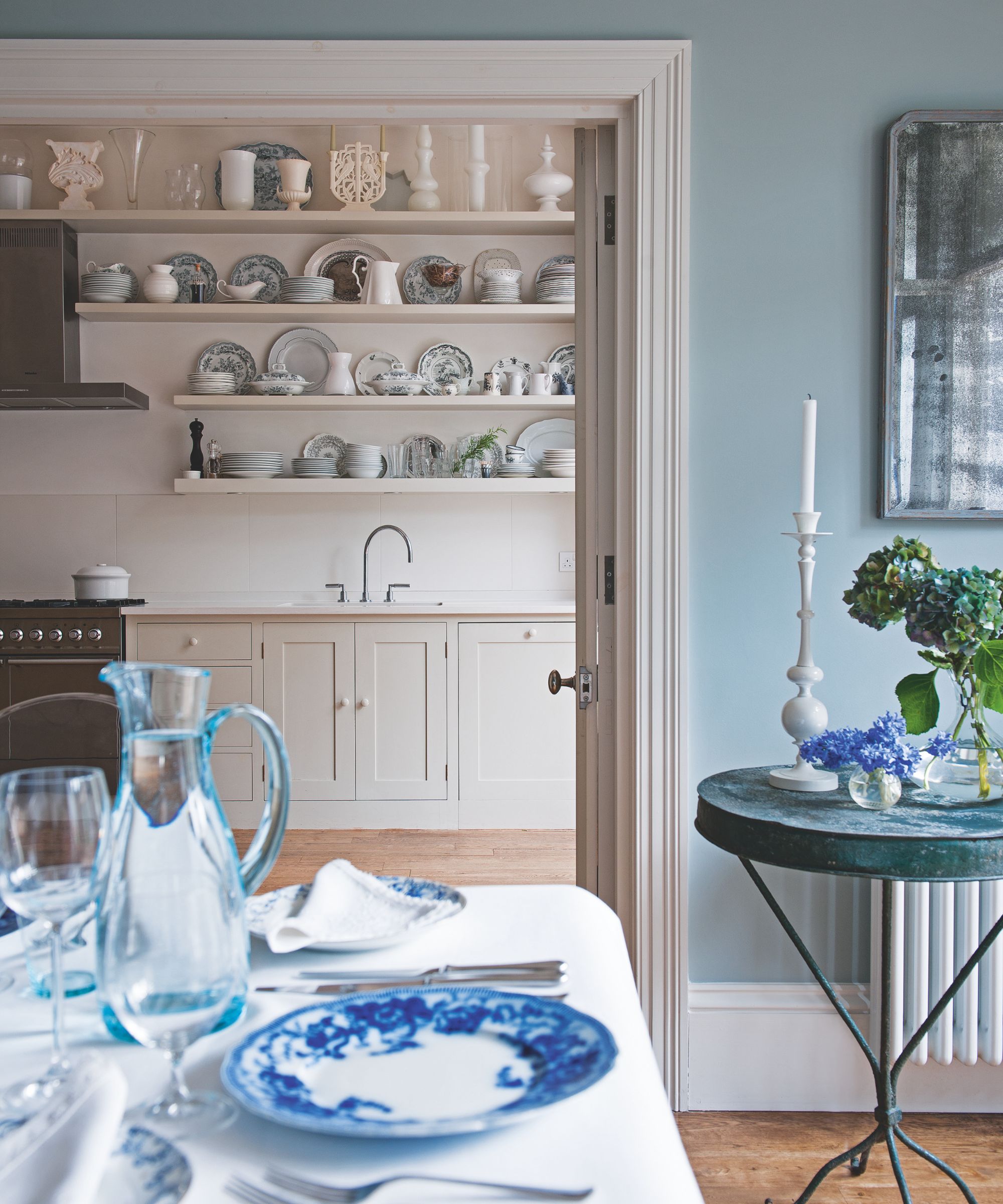 Dining room with blue and white laid table looking into kitchen with shelves of china and ceramics