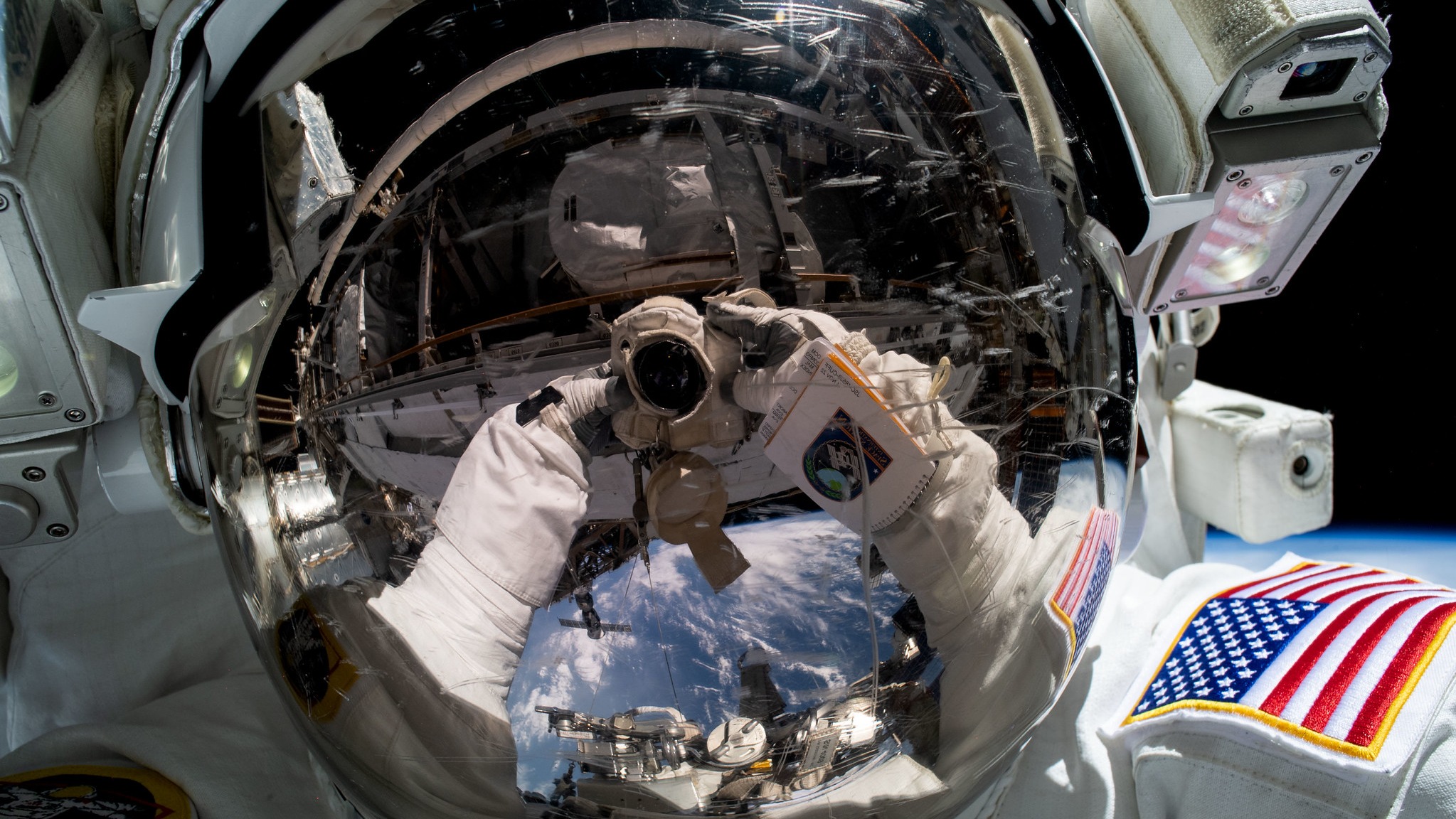 view of a spacesuit helmet up close with the reflection of two arms. in the reflection a camera is visible in the gloved hands, taking a picture. the black of space and a bit of the curve of earth is visible behind the helmet