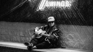 A black-and-white shot of Marcus King sitting by a pool playing a Gibson acoustic. Mood Swings, the title of his new album, is lit up in neon above him.