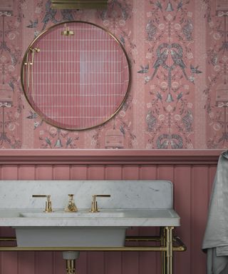 A pink bathroom with pink floral wallpaper with blue flowers, a gold mirror reflecting pink tiles, and a white marble sink with gold taps