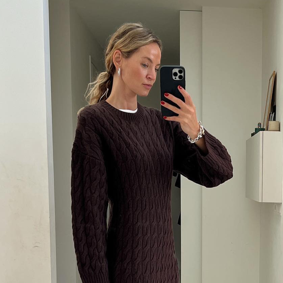 Anouk Yve is in a brown sweater dress, brown heeled boots, and a curved brown shoulder bag.