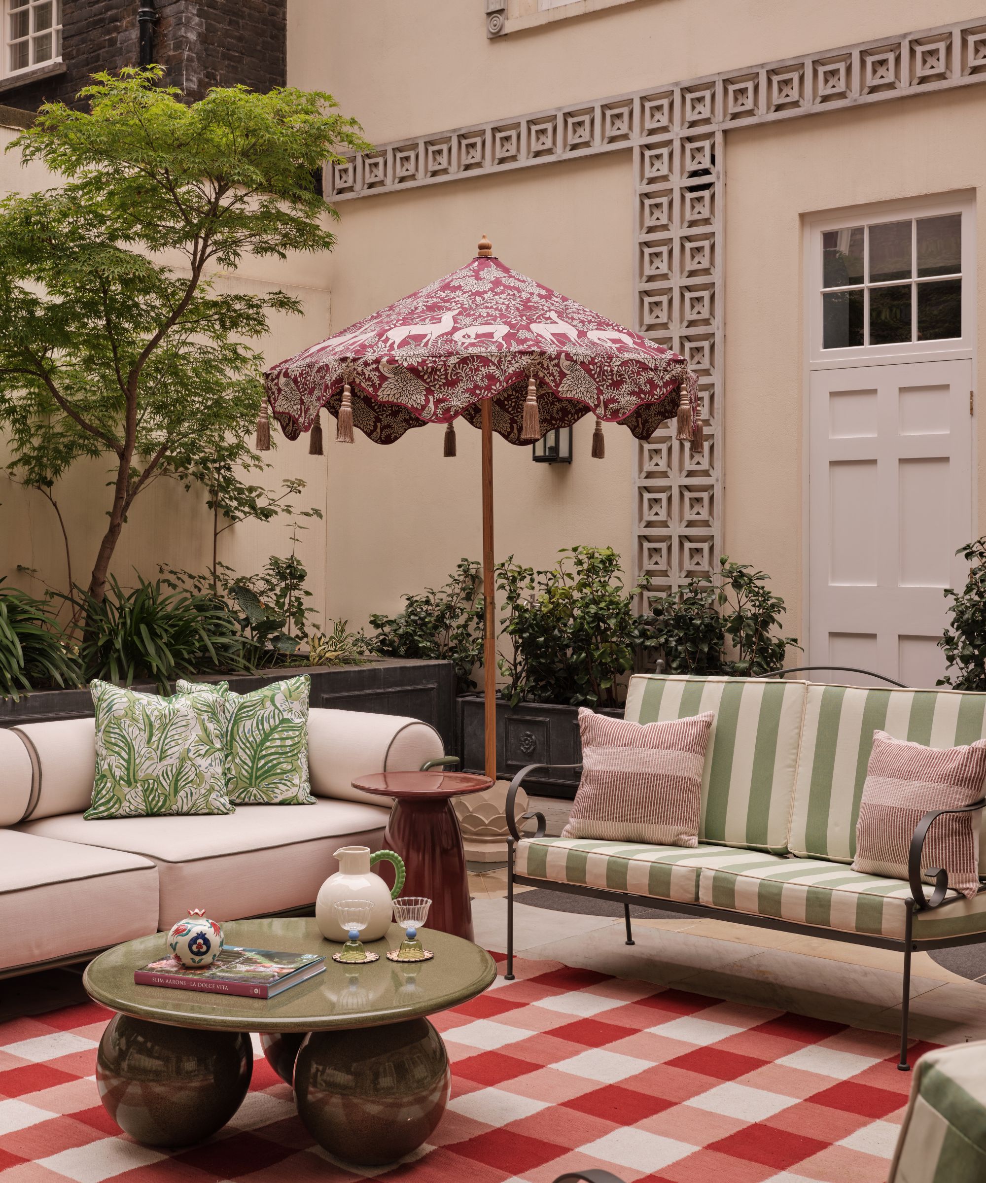 Garden with plants growing on the wall, a large red gingham rug with a large cream sofa on top, green metal armchairs with striped green cushions, and a marble green round coffee table at the centre.