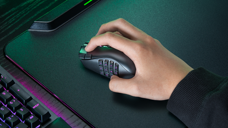 A hand holding a Razer Naga V3 Pro mouse on a mouse mat next to a Razer keyboard