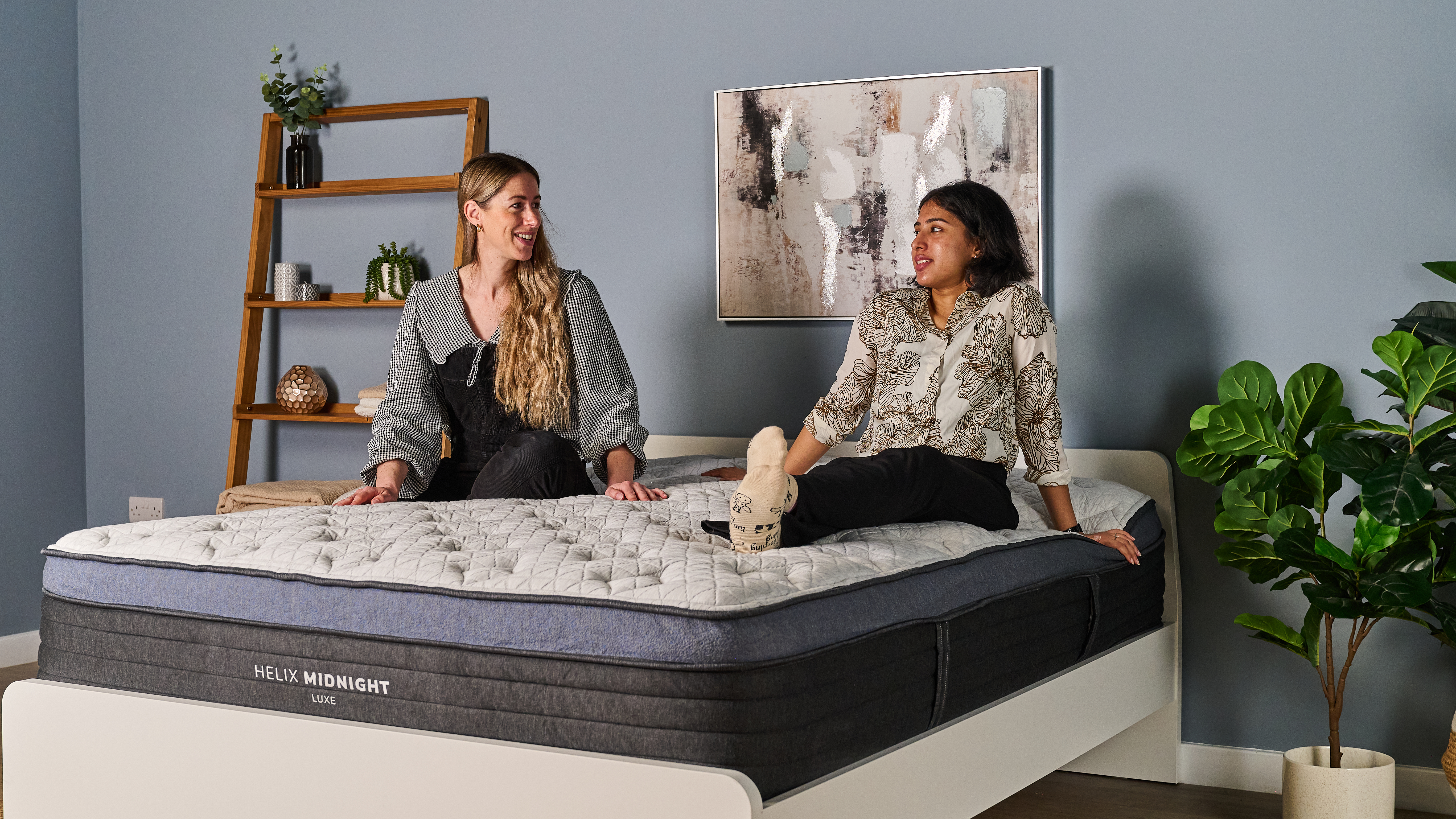 The image shows two mattress testers sat on top of the Helix Midnight Luxe, which is placed on a white bed frame in a blue bedroom