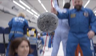 A plush ball resembling a cratered moon floats in the cabin of an airplane during a parabolic flight.