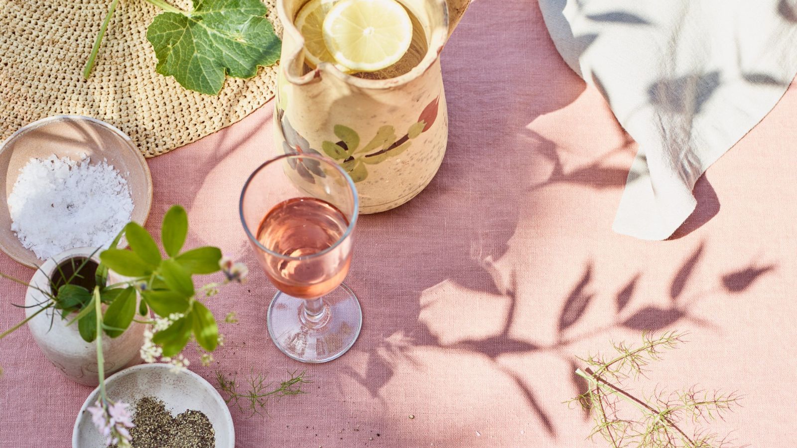 Table with pink tablecloth, jug of lemon water, glass of rose, salt and pepper pinch pots, rattan placemat and some greenery