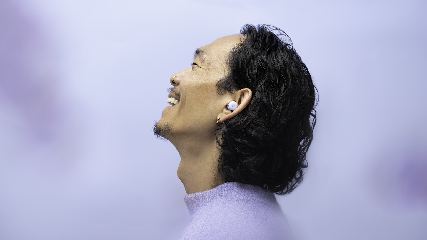A smiling man in a lilac jumper against a lilac background wearing a pair of Google Pixel Buds 2a in lilac, although Google calls that color Iris