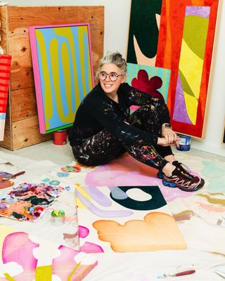 A woman dressed in black clothes covered in paint sits in a colorful studio filled with just as colorful canvases.