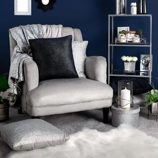 A light grey armchair with grey cushions and a throw, a white fluffy rug and silver ornaments on glass shelves