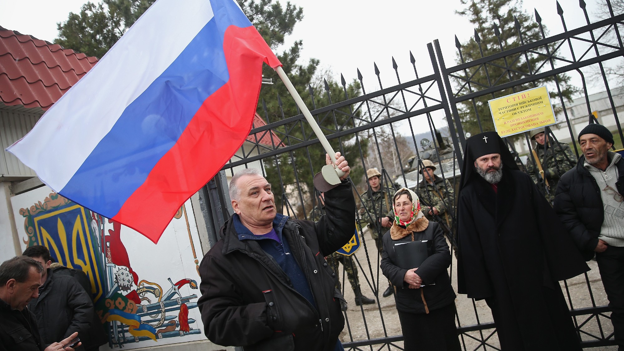 A man waves a Russian flag next to the gate of a Ukrainian base with Ukrainian soldiers watching on