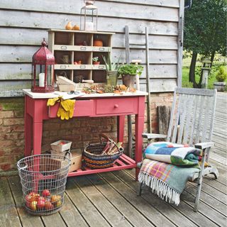 Pink potting bench on wooden decking area next to wooden deck chair and metal basket of apples