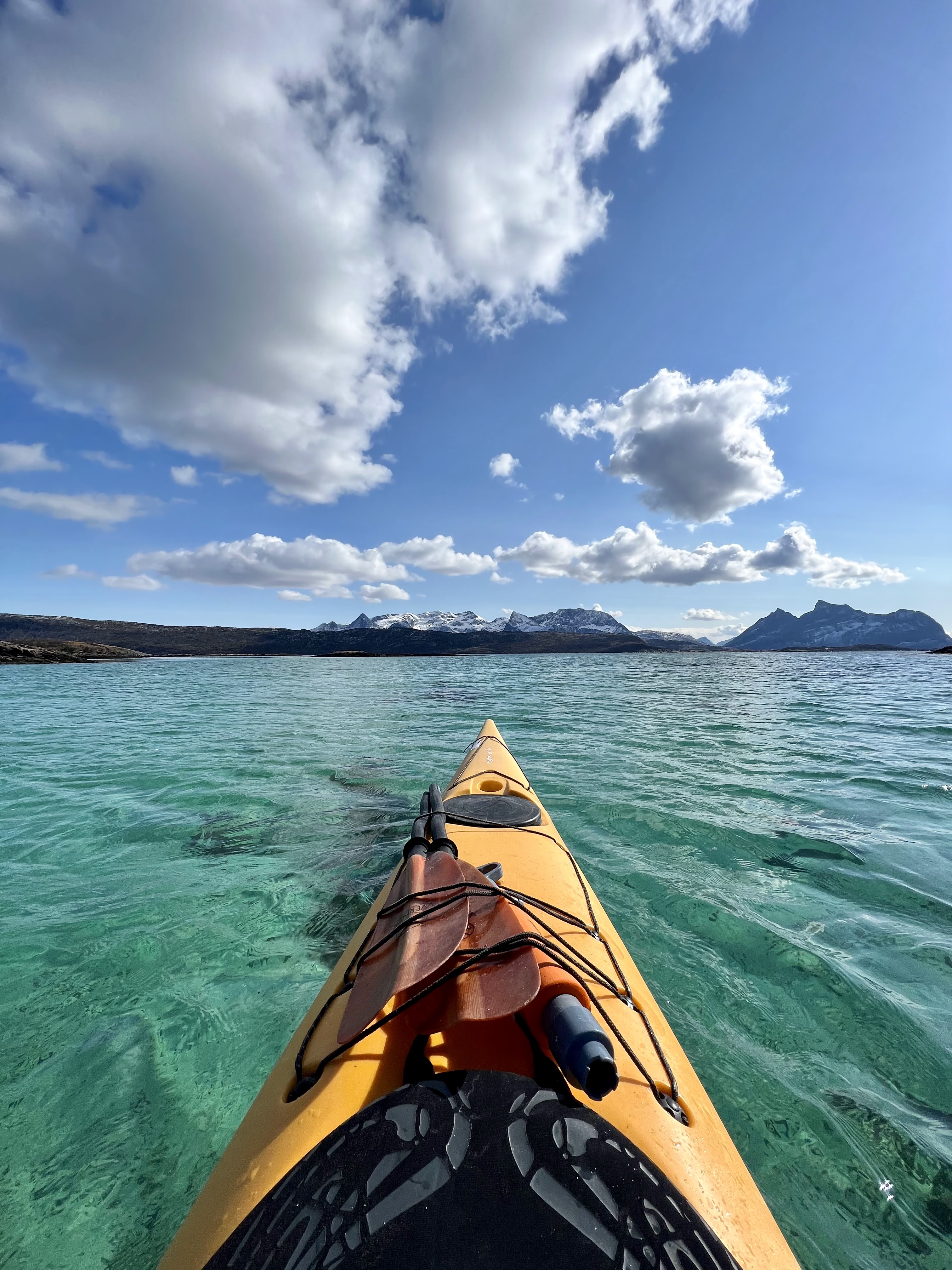 View of the nose of a kayak in a fjord in Lofoten Island in blue/green crystal clear water with snowy mountains in the background in a sunny day with a few clouds in the sky.