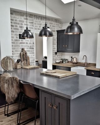 A grey kitchen with three pendant ceiling lights over island, grey brick exposed wall and bar stools