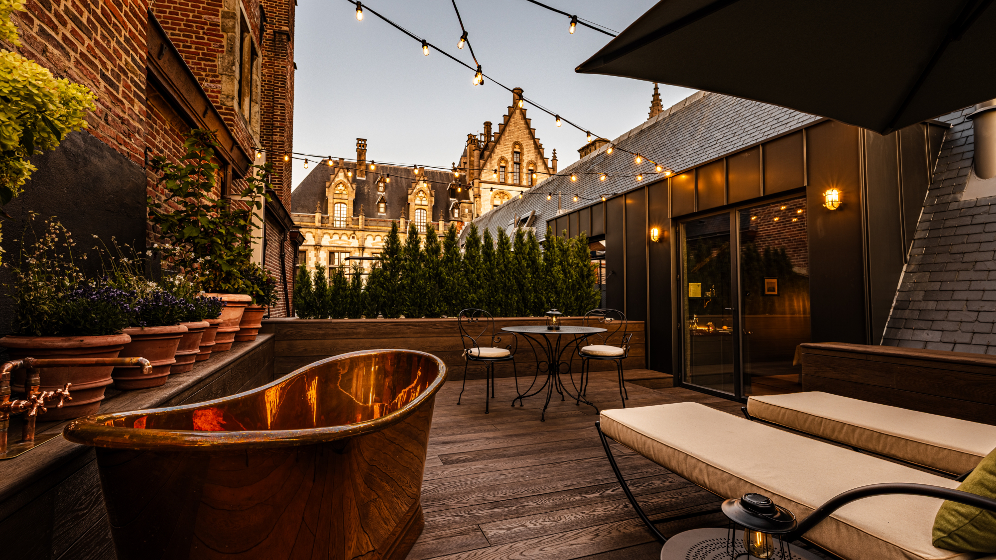 Roof terrace at night, with copper bath, sunloungers, string lights and view of a gabled building, at 1898 Porter&amp;rsquo;s House, Ghent, Belgium
