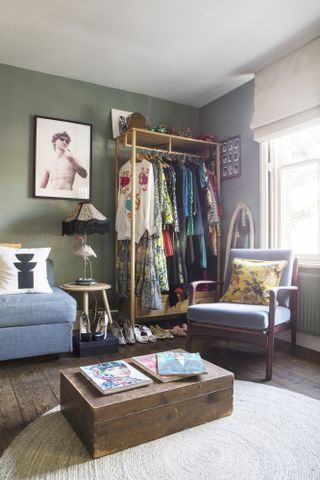 Area of the home office with open wardrobe filled with clothes and shoes, seating area with a sofa and an armchair, a coffee table topped with fashion magazines and framed photograph against an olive green wall