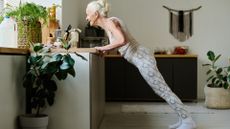 woman sideways to the camera doing a pushup against a kitchen counter in a home kitchen setting with plants in front and behind.