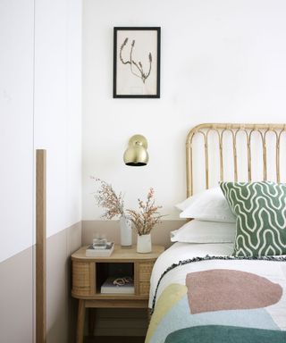 Relaxed bedroom with white painted walls, textured, bamboo style bedframe, white bedding, patterned cushion and pastel throw, brass wall light, wall art mounted above, wooden side table, taupe painted trim on wall