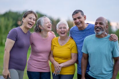 A small group of mature adults stands together as they pose for a portrait while taking their fitness outdoors. They are dressed in athletic wear and are smiling.