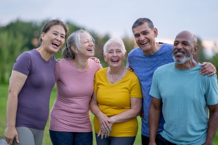 A small group of mature adults stands together as they pose for a portrait while taking their fitness outdoors. They are dressed in athletic wear and are smiling.