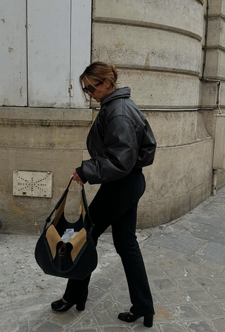 what to wear in 30-degree temperatures, shown in a photo of a woman walking down the sidewalk wearing a leather bomber jacket with jeans, pumps, and a hobo bag