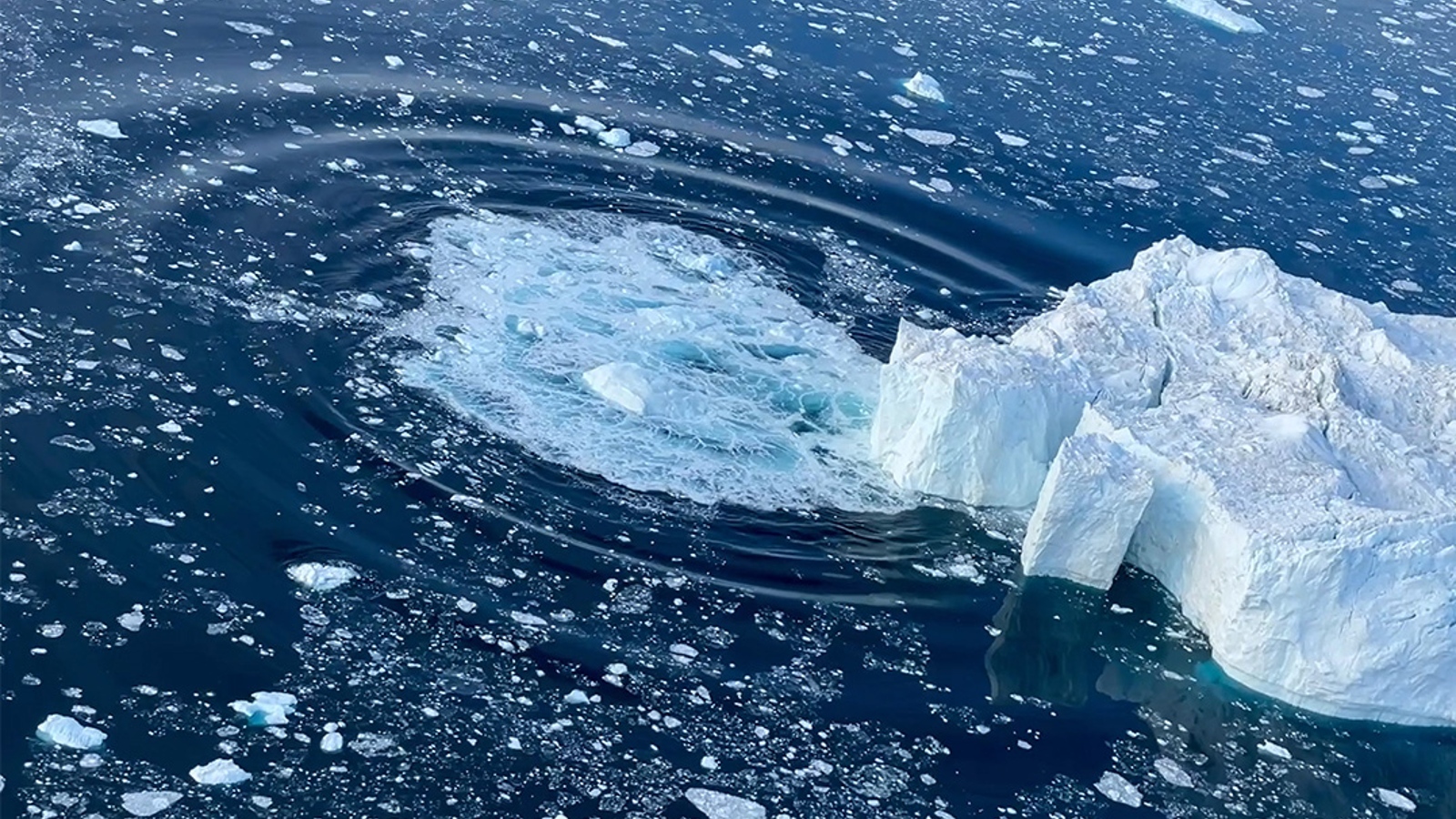 A photo of a large chunk of ice falling into the sea from a glacier, creating a circular wave