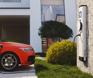 An electric car charging point mounted to a wall at a home charging a red car on a driveway. A modern home in the background