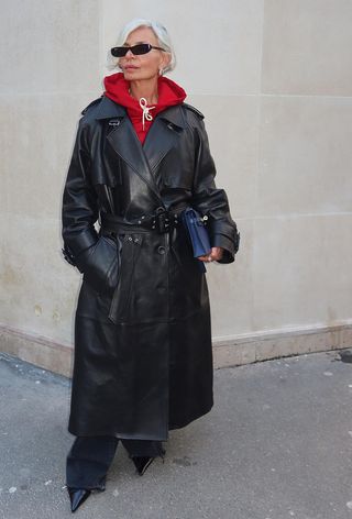 A woman standing outside wearing a red hoodie under a black leather trench coat with wide-leg dark-wash jeans and pointed black pumps.