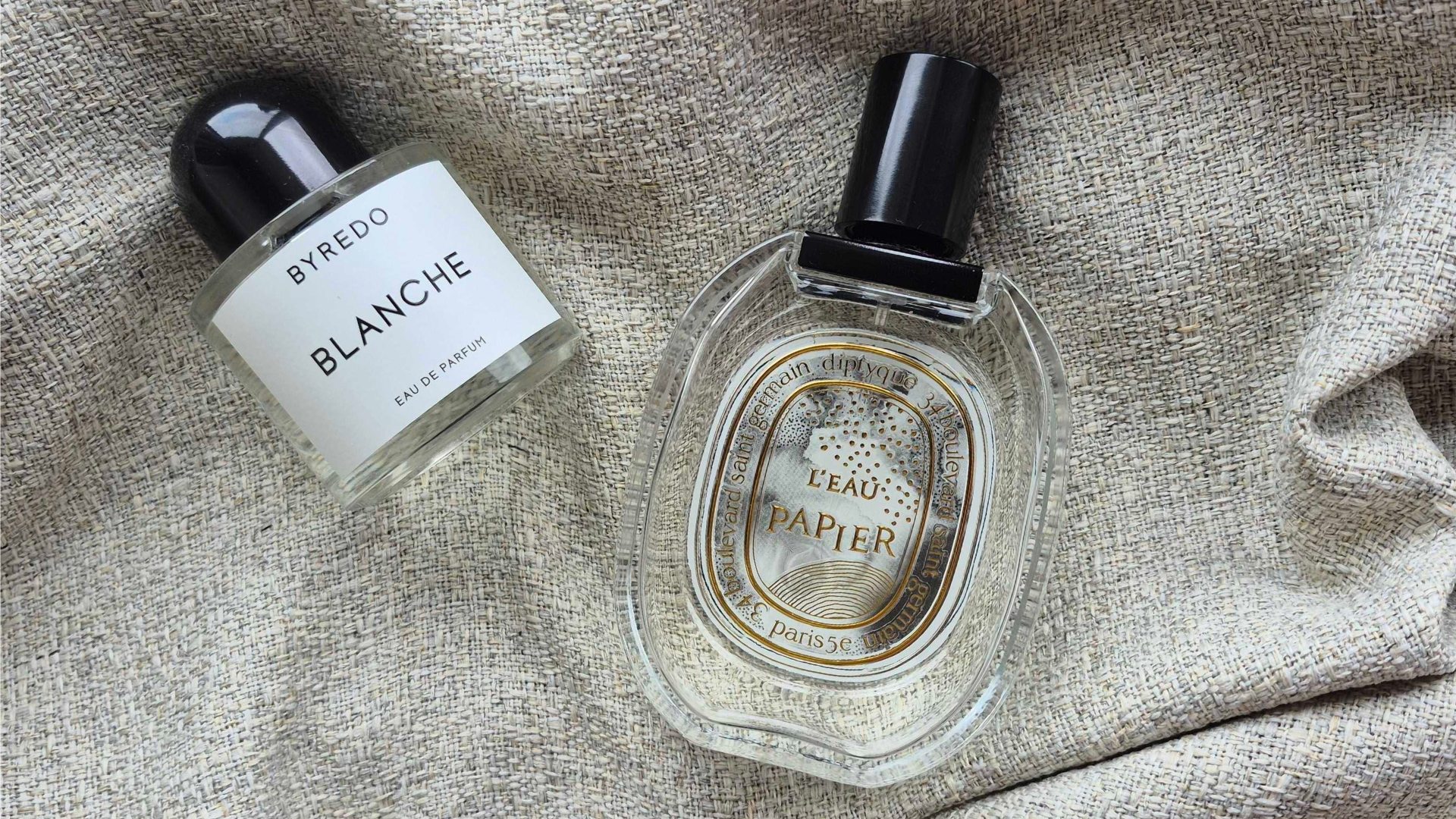 A picture of Byredo's Blanche and Diptyque's L'eau Papier perfumes laid out on a grey linen fabric background - photographed by Digital Beauty Writer, Naomi Jamieson