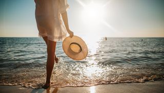 summer, summer solstice, Close-up of young woman in white sun dress and with hat in hand walking alone on sandy beach at summer sunset, splashing water in sea shallow