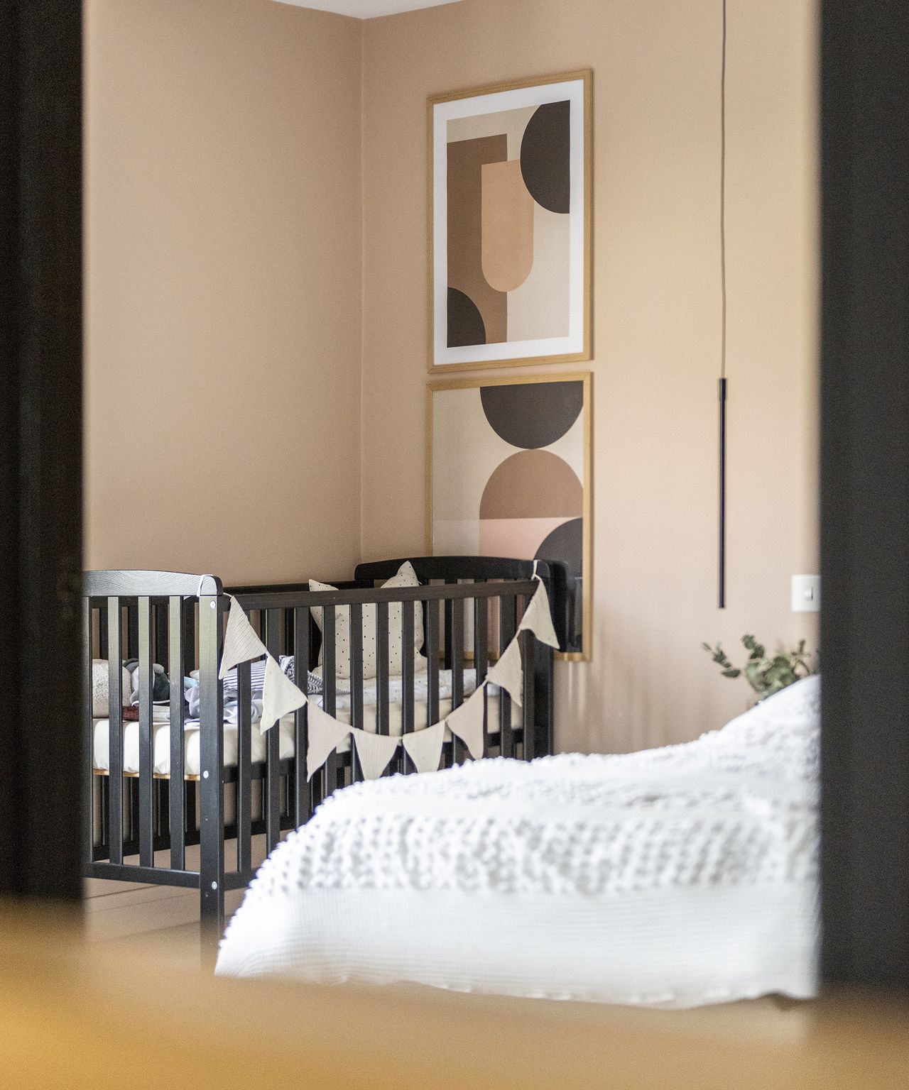 A black child's cot in the corner of an adult's bedroom. The walls are a soft plaster-pink with matching wall art in frames
