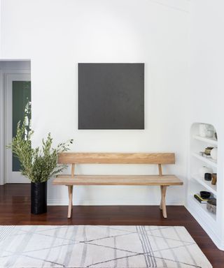 White entryway with wooden bench, gray square artwork, niche shelves, plant, rug on dark wood floor