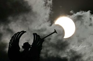 the sun on the right looks like a "bite" has been taken out of it as the moon moves over the solar disk, the sun appears as a crescent shape. to the left is a statue of an angel with wings and a large trumpet like instrument, it is pointing it at the sun.