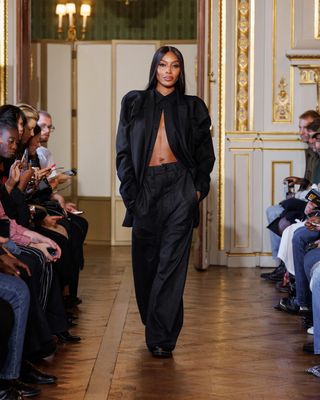 Naomi Campbell walking in Torishéju Dumi Spring 2024 show in a black blouse, black pants, and black shoes.