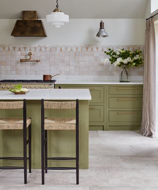 green and nude kitchen with green cabinetry and island, white marble countertops, tiled floor, nude backsplash, brass hardware, pendant lights, rattan bar stools, drapes