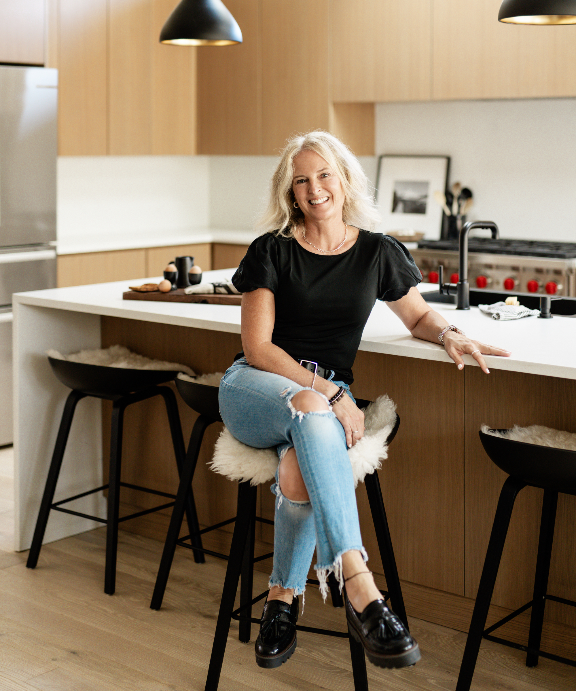 image of Shelagh Conway Triple Heart Design, blond woman wearing black top and jeans in a wood and white kitchen