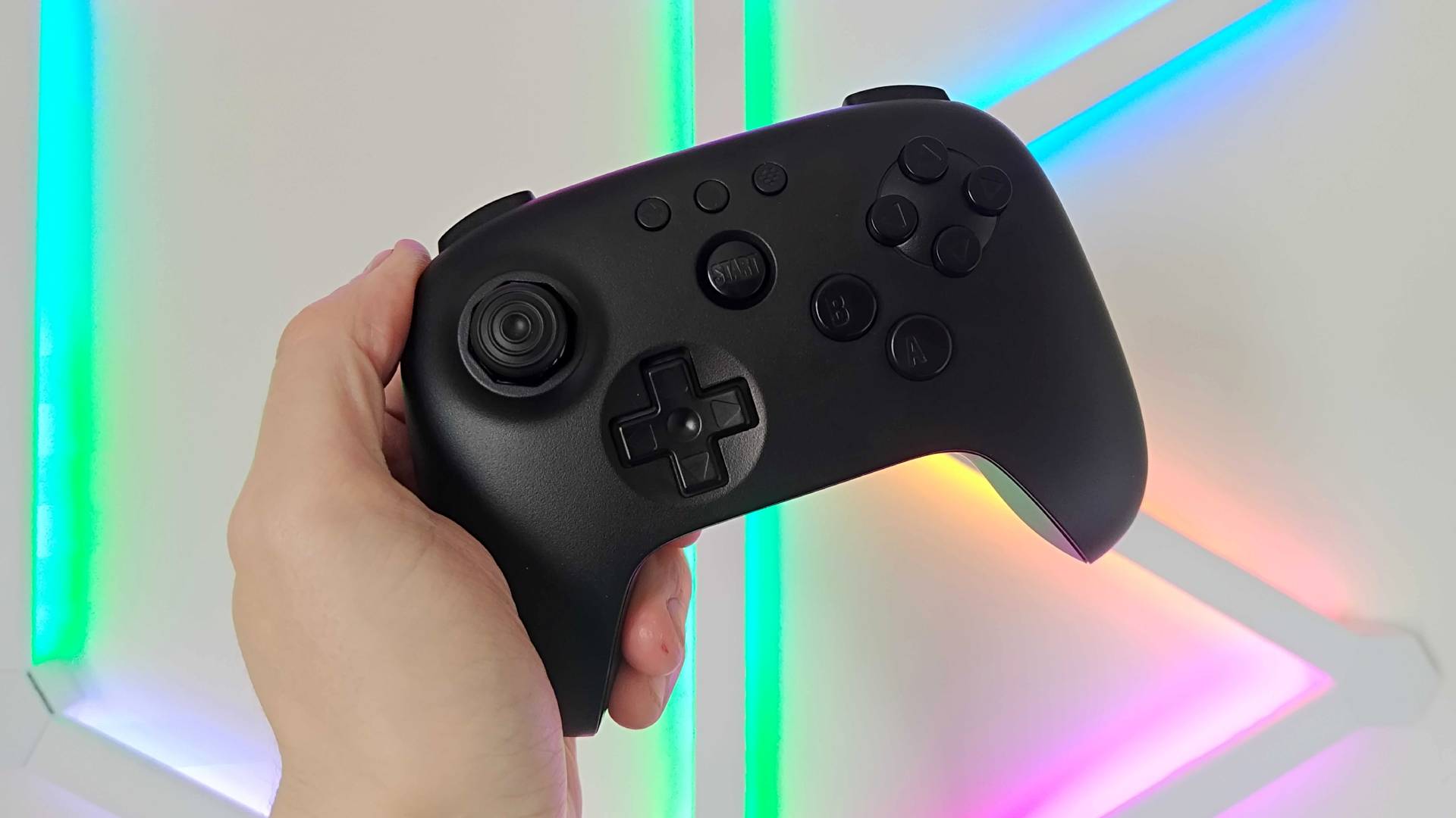 Hand holding 8Bitdo N64 controller with RGB lights in backdrop on white wall.